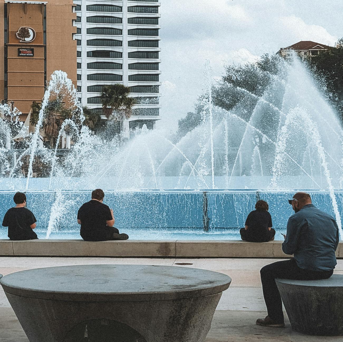 Adults relaxing by a fountain in downtown Jacksonville with a cityscape background.