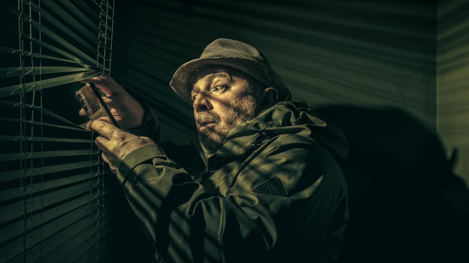 A man in a hat suspiciously peering through blinds, holding a phone in shadowy light.