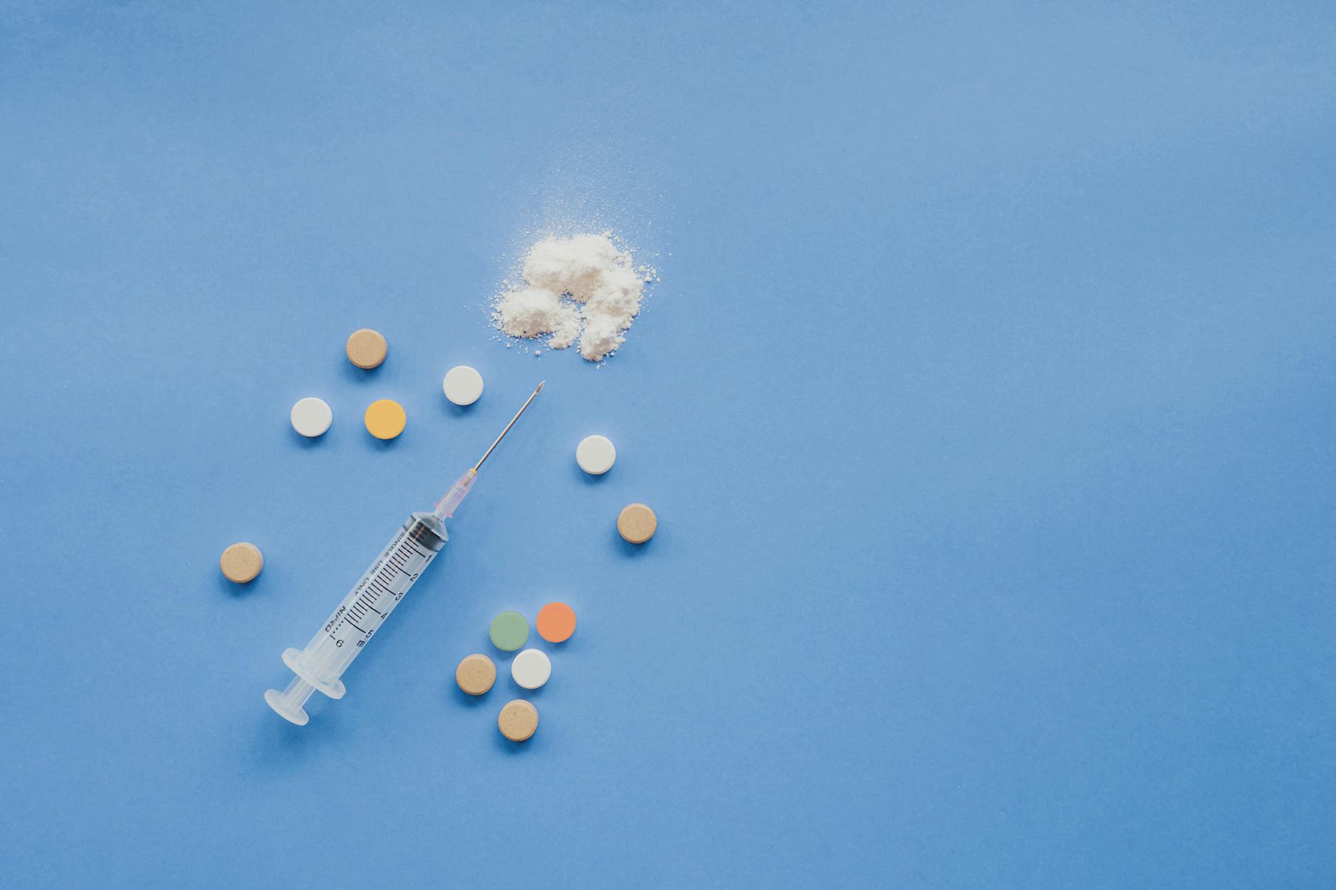 From above of syringe with multicolored pills and powder placed on blue background